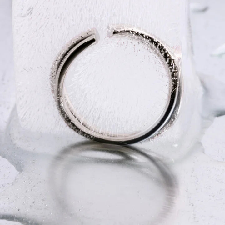 Bague ondulée Anomalie, graphique et ouverte, en métal satiné argenté, présentée en nature morte sur un fond de glace.