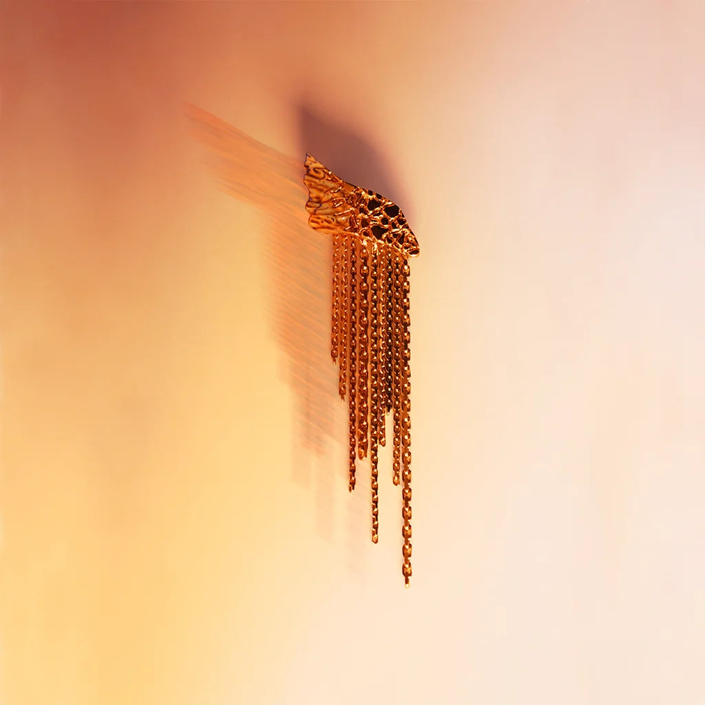Une boucle d’oreille originale créateur Trouble en vieux doré en nature morte sur un fond de couleur chaudes.