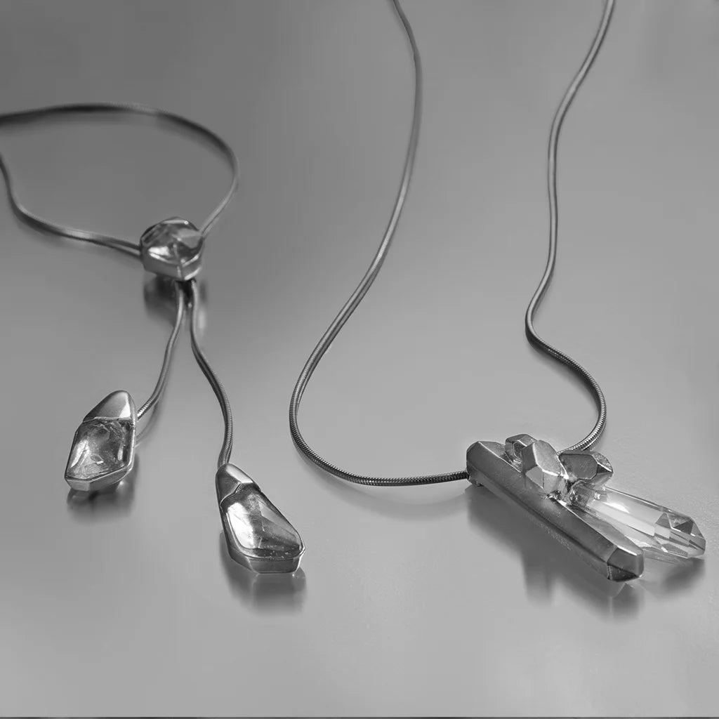 Pendentif cristal de roche et collier cravate Turbulence en métal argenté, posés avec souplesse sur fond gris.