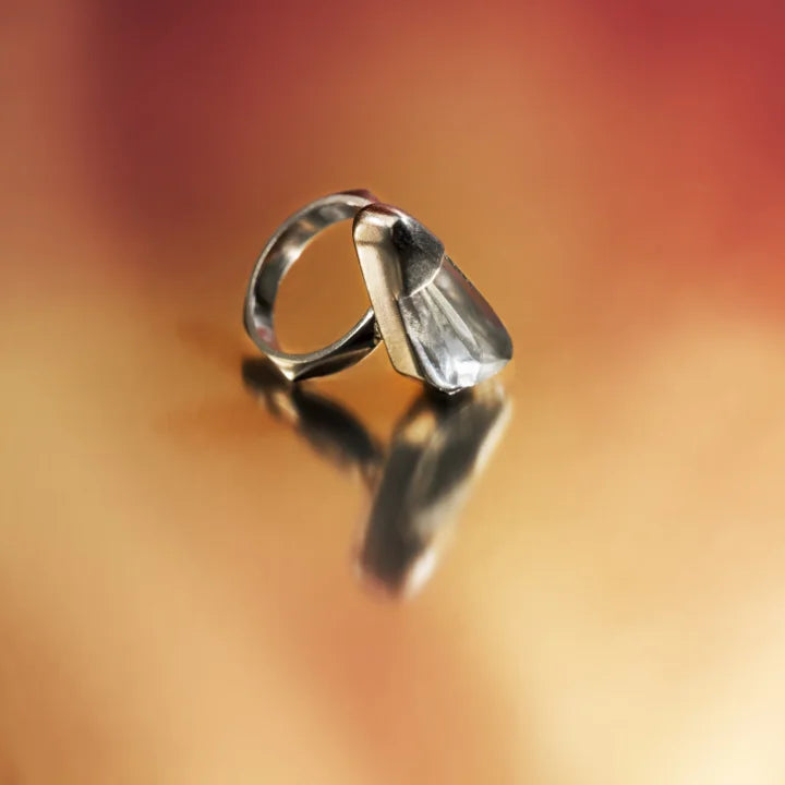 Une bague cristal de roche Turbulence en métal satiné doré posée de manière décorative sur un fond aux couleurs chaudes.