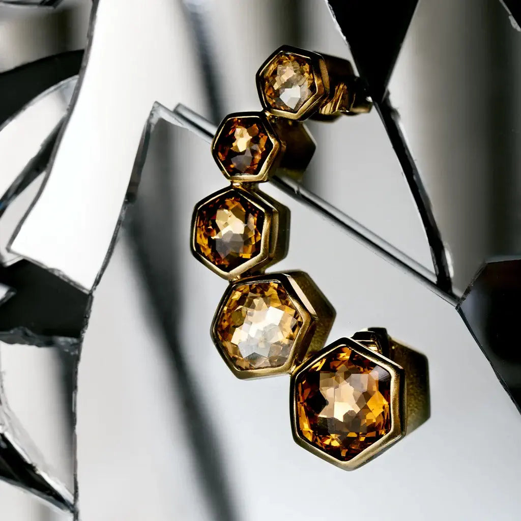 Une boucle d'oreille strass Alma en métal satin doré jaune et camel posé en nature morte sur des verres cassés.