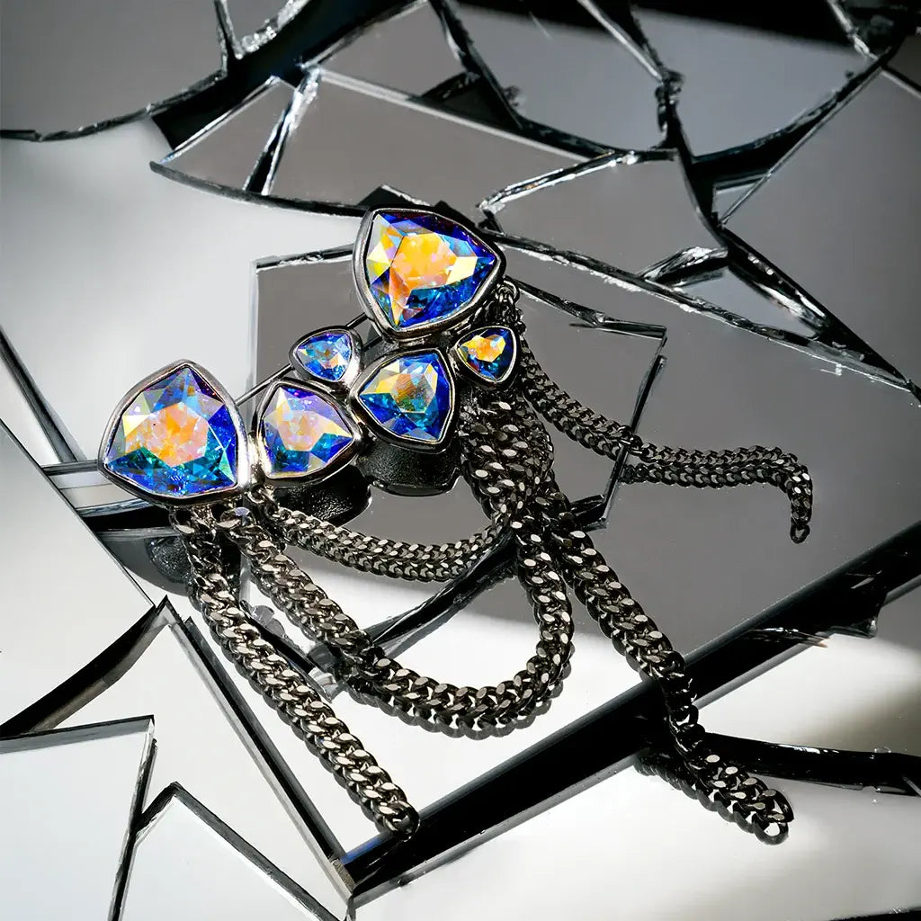 Une grande broche en métal satin argenté et aurore boréale, présentée en nature morte sur des verres cassés.