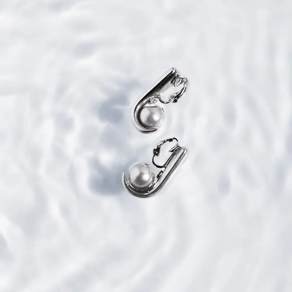 Des boucles d’oreilles Noe à clip en métal satin argenté et perles en cristal laqué gris, posées sur fond blanc neige.