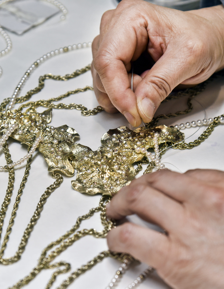Mains assemblant un collier en métal doré et perles, travail minutieux de création de bijou sur une table d’atelier.