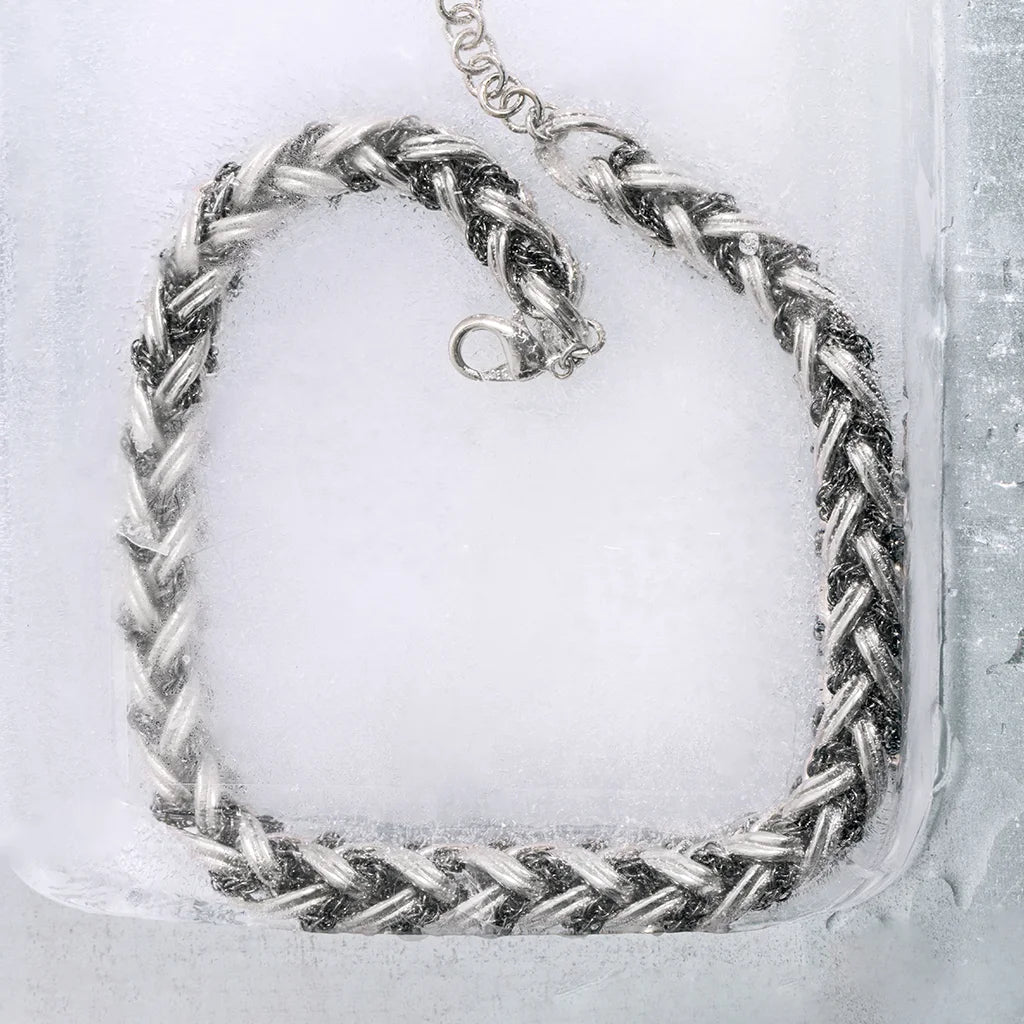 Collier tressé Anomalie en métal bicolore argenté et ruthénium posé sur un fond de glace, créant un effet de fluidité.