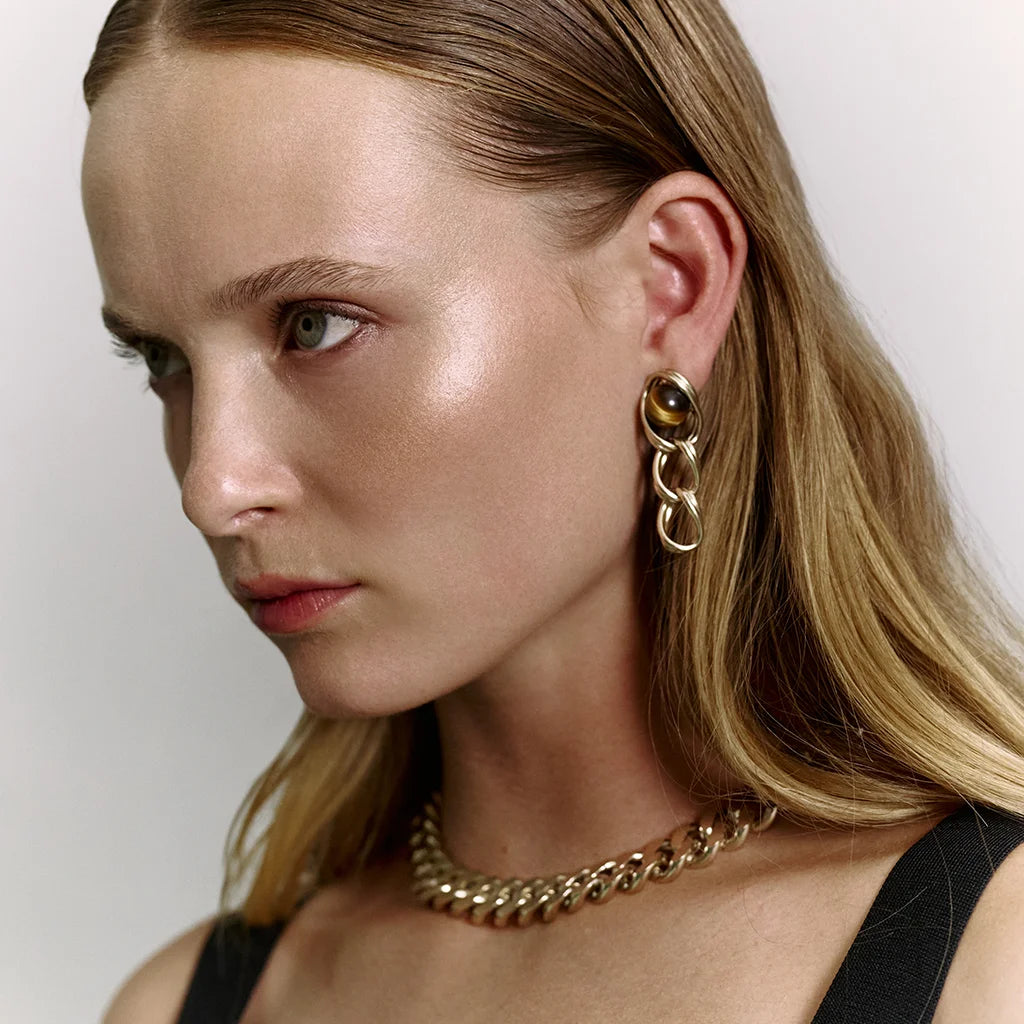 Femme portant des boucles d’oreilles perles pendantes Anomalie, en métal satiné doré et œil de tigre avec un collier.