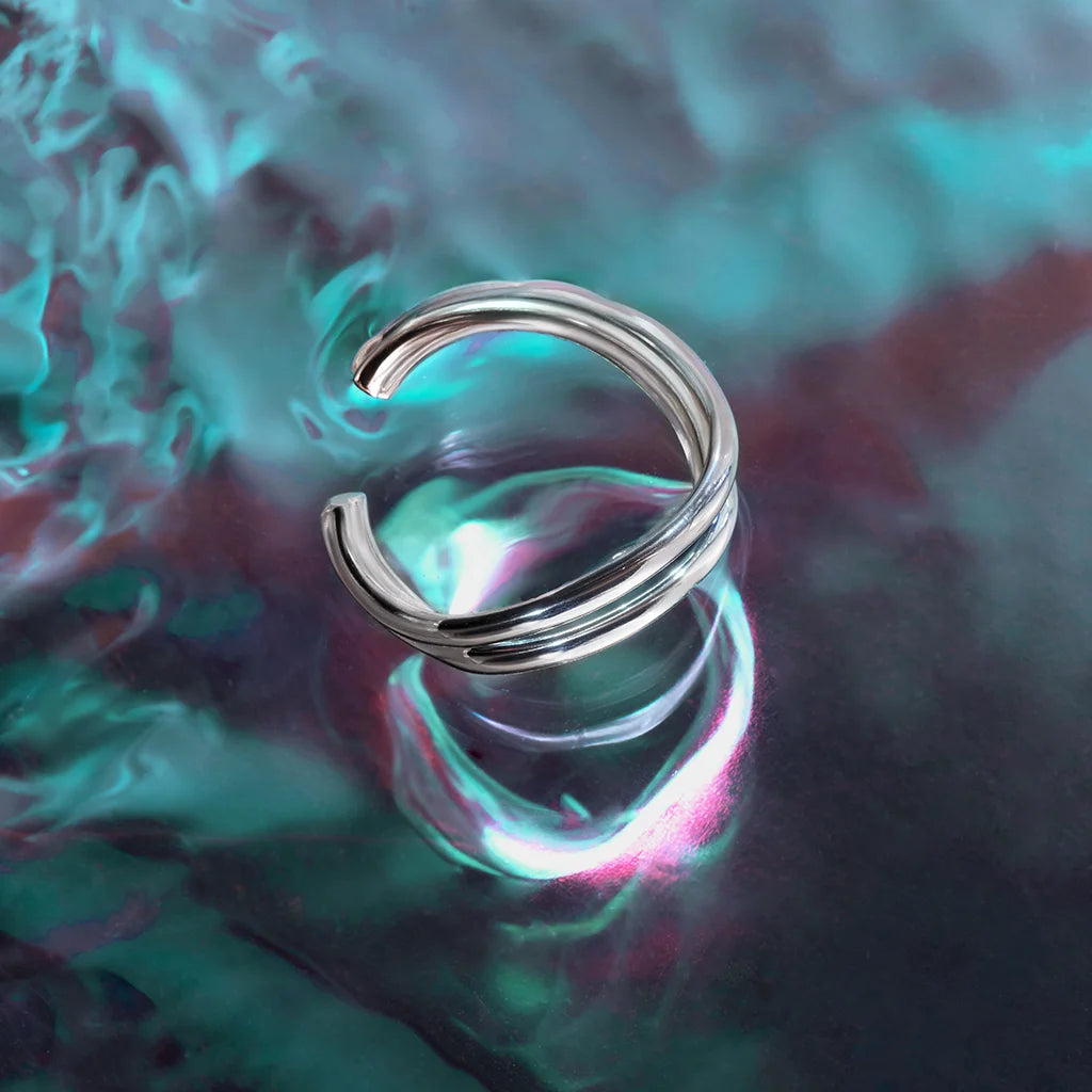 Une bague ondulée Anomalie, graphique et ouverte, en métal satiné argenté, présentée en nature morte sur fond coloré.