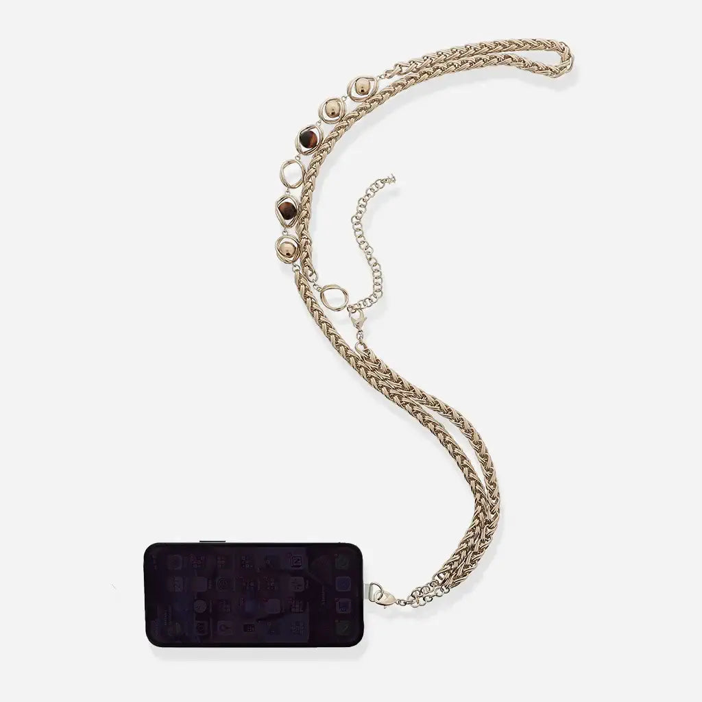 Une bandoulière de téléphone Anomalie en métal satiné doré, composée d’une chaîne tressée aux maillons graphiques et perles naturelles.