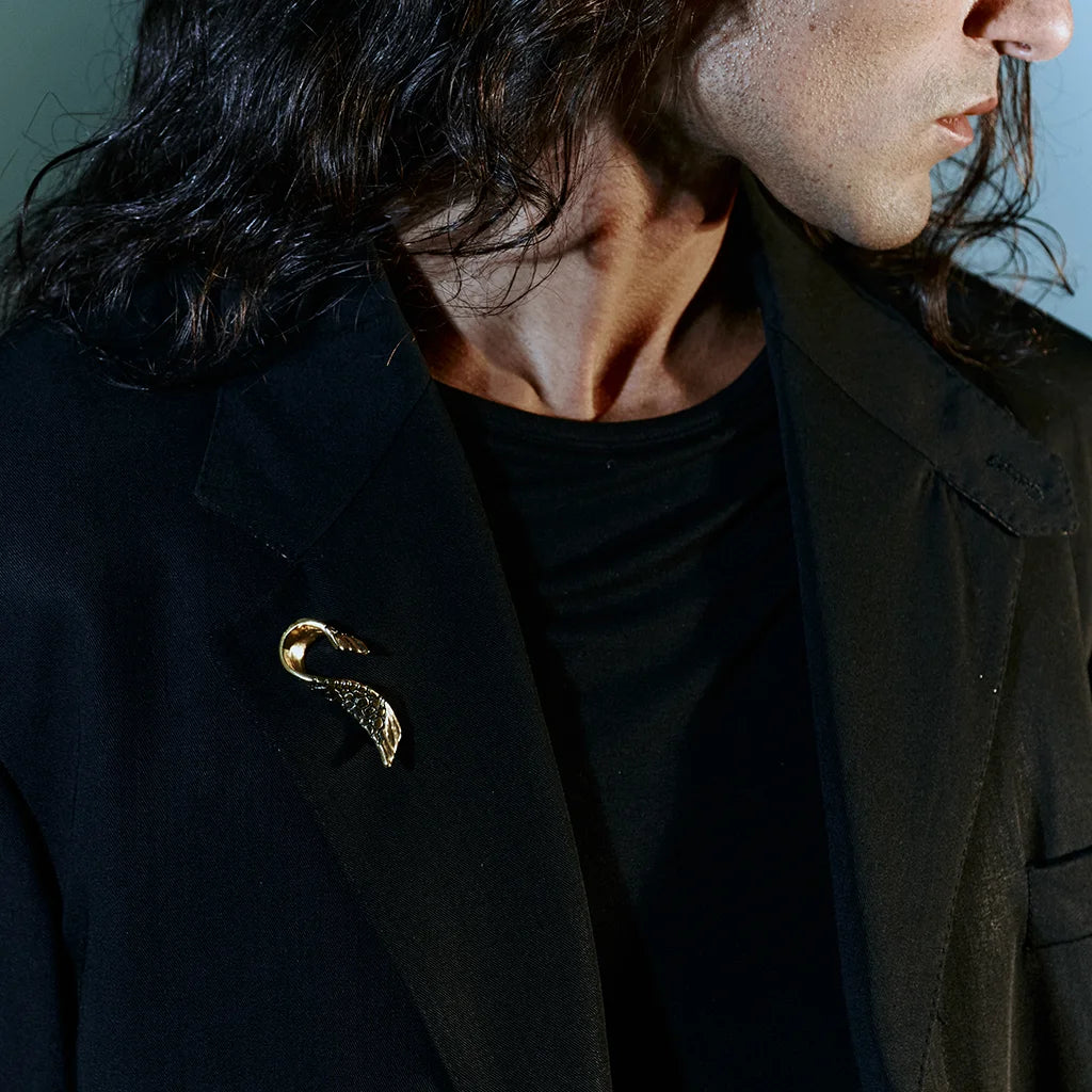 Une broche fibule Trouble en métal texturé vieux doré, au design d’aile torsadée, portée sur le blazer d’un homme.