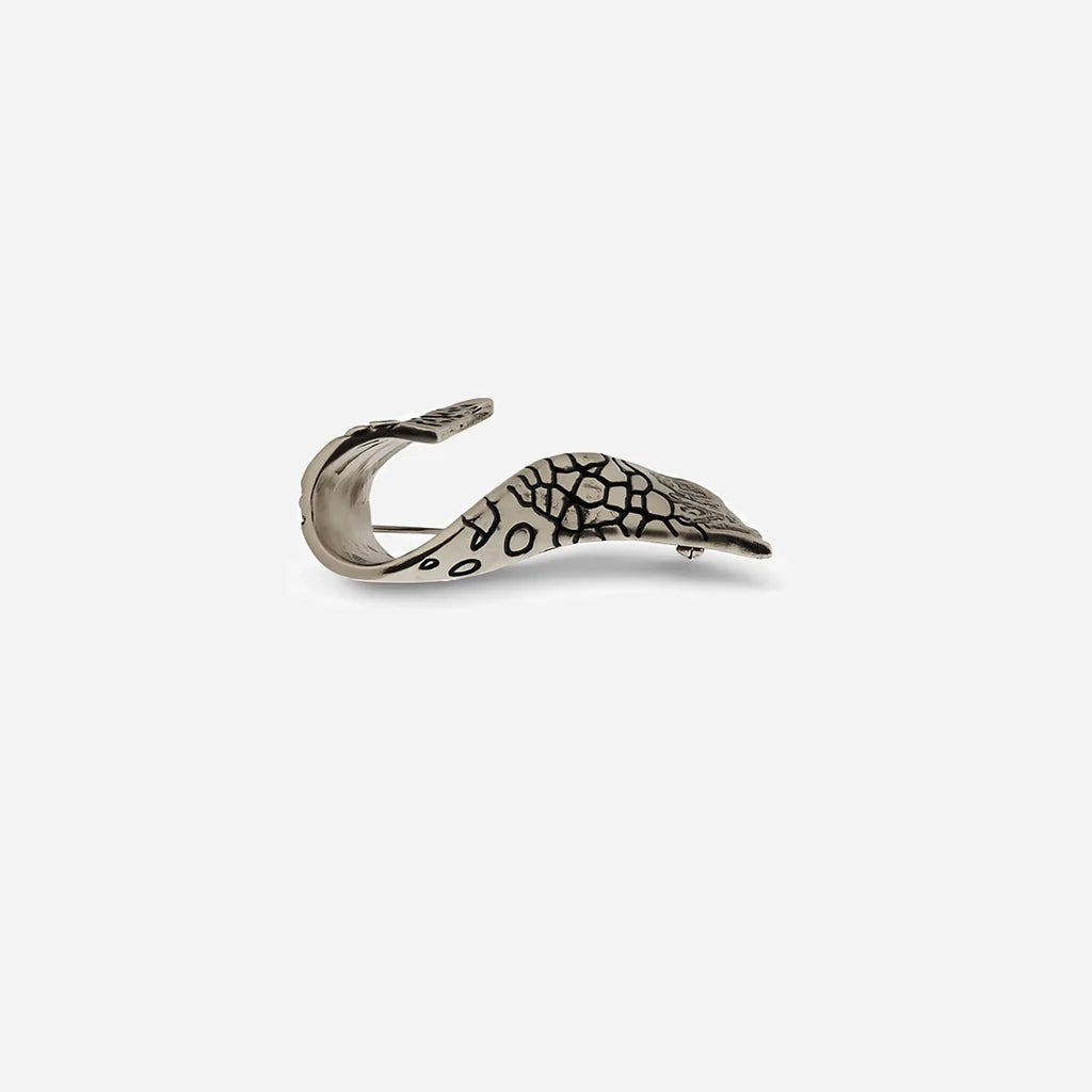 Une broche fibule Trouble en métal vieil argenté, au design d’aile torsadée avec un travail de texture prononcé.