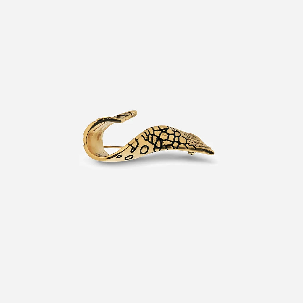 Une broche fibule Trouble en métal vieux doré, au design d’aile torsadée avec un travail de texture prononcé.