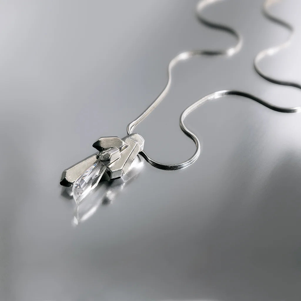 Nature morte composée d'un pendentif cristal de roche Turbulence avec chaine serpent souple en métal argenté posé sur un fond aluminium.