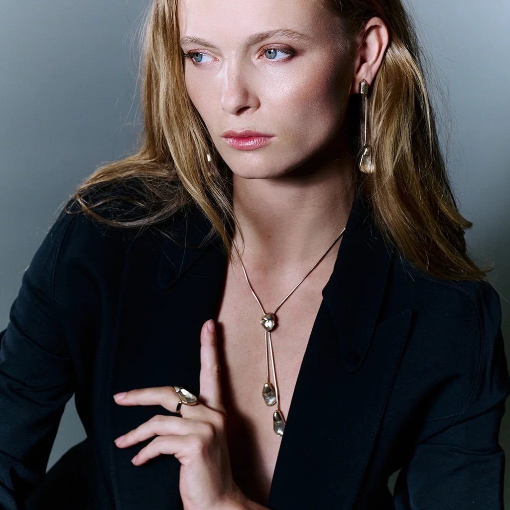 Femme portant un collier cravate, des boucles d'oreilles cristal de roche et une bague cristal de roche Turbulence en métal doré.