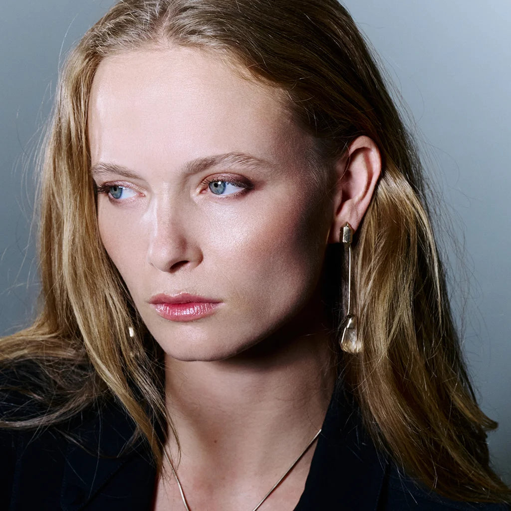 Une femme portant des boucles d'oreilles cristal de roche Turbulence en métal doré, à la silhouette longiligne et graphique.