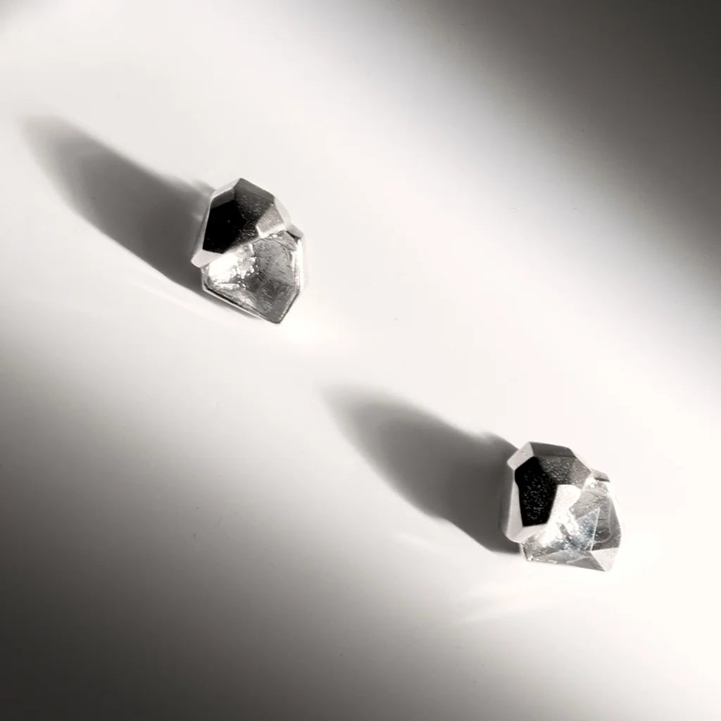 Des puces d'oreilles Turbulence en métal argenté sur fond gris, compactes et structurées, mettant en valeur l'éclat brut.