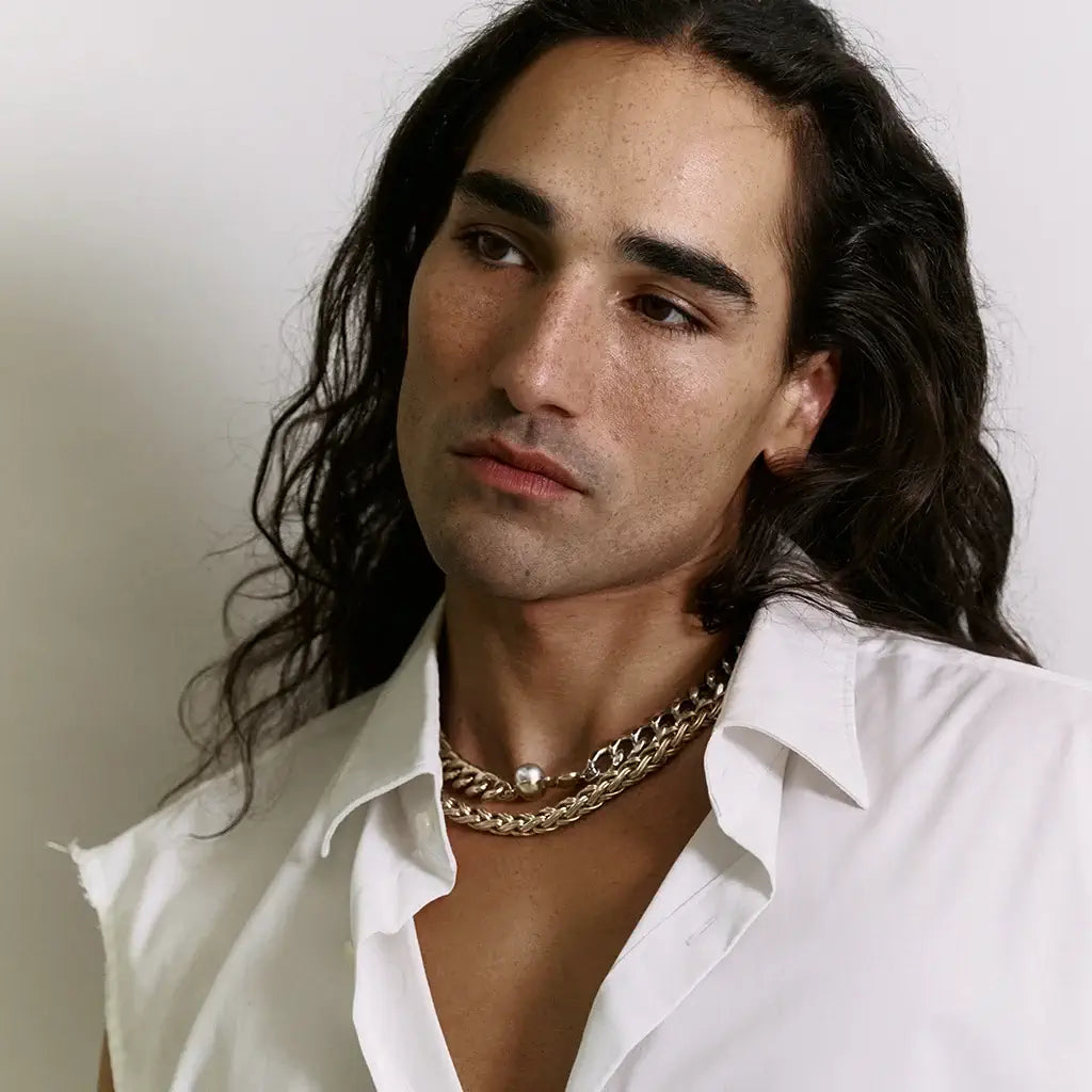 Un homme portant deux colliers : un collier Fleur Chloé argenté et doré brillant avec perle grise, et un autre collier.