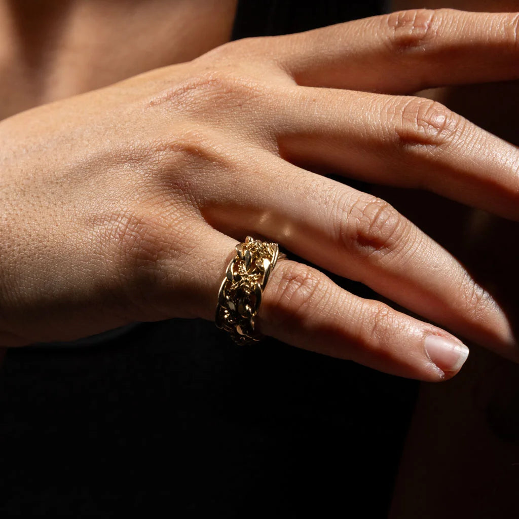Bague chaîne sculpturale en métal doré portée au doigt, photographiée en gros plan sur une main dans une lumière chaude et contrastée.
