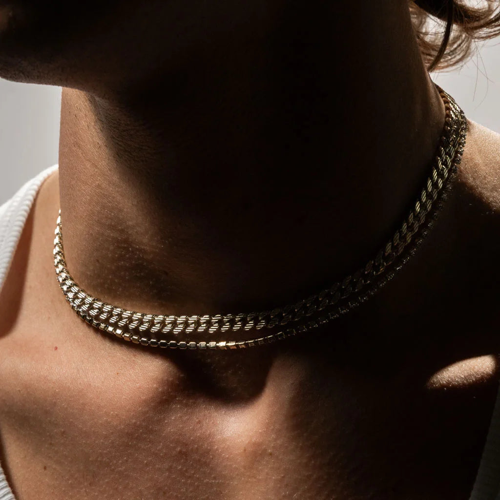 Collier chaîne double en métal doré porté ras du cou, photographié en gros plan dans une lumière chaude et contrastée avec reflets métalliques.
